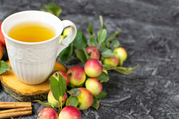 A white mug with tea on a wooden stand, ripe red apples on a branch, cinnamon sticks on a black background. Composition. Space for text.