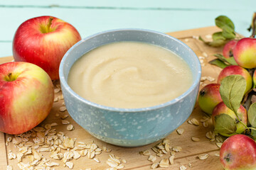 Oatmeal porridge for the baby from ground cereals in a blue bowl, red ripe apples on a wooden board on a blue background close-up. Baby nutrition, the first complementary feeding of a child.