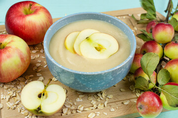 Baby's first complementary food, baby nutrition. Oatmeal porridge for the baby from ground cereals in a blue bowl, red ripe apples on a wooden board close-up.