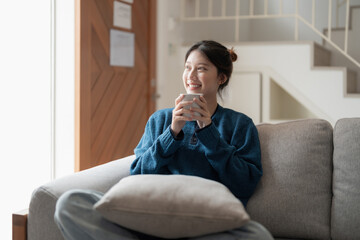 Happy young asian woman sitting on a couch with a cup of coffee at home