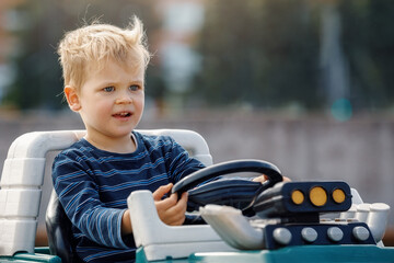 Cute little boy driving big electric toy car with steering wheel and having fun outdoors.