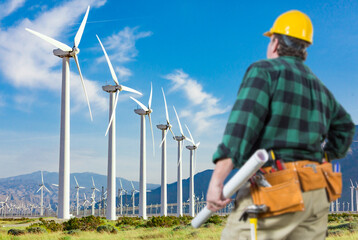 Male Contractor Wearing Tool Belt and Hard Hat Facing Alternative Energy Wind Turbines.