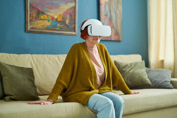 Mature woman in vr goggles sitting on sofa in living room and watching virtual reality