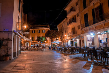 Mallorca, Spain. April 27, 2022. Illuminated outdoor restaurants and cafe at townsquare. Hotel buildings with place setting in city against sky. View of town during night.