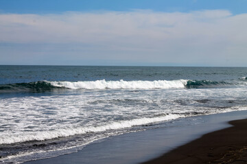 The coast of the Pacific Ocean is covered with black volcanic sand. Long turquoise surf waves roll and foam. Blue sky. Kamchatka. Khalaktyrsky beach. High quality photo