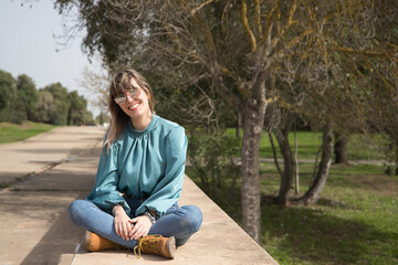 Fototapeta premium Young, pretty, blonde woman in green shirt, jeans and mirrored sunglasses, sitting smiling in a park. Concept warmth, spring, summer, vacation.