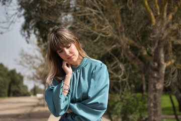 Portrait of young, beautiful, blonde woman in green shirt, with hand resting on chin sitting seriously in a park. Concept depression, problems, seriousness, concern.