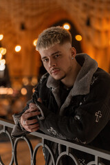 Fashionable young man hipster with hairstyle in trendy stylish black jacket stands in the mall