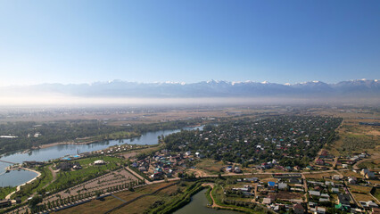 Landscape with a view of lakes, beautiful houses and snowy mountains. There are cars on the road. Green streets among the houses. Tall trees and fields. A quiet little town. Kazakhstan, Almaty