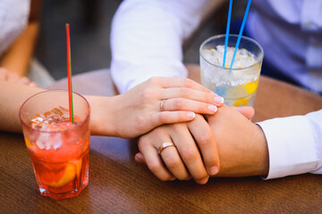 Fototapeta premium The bride’s hand is on the groom’s hand. Couple sitting at the table, close-up hands with wedding rings and cold drinks