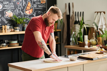 Modern Caucasian man wearing casual clothes standing in loft kitchen kneading and flattening dough...