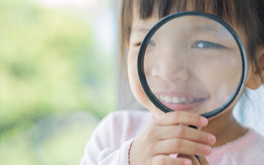 Portrait adorable Asian little girl holding magnifying glass to investigate, smiling with...
