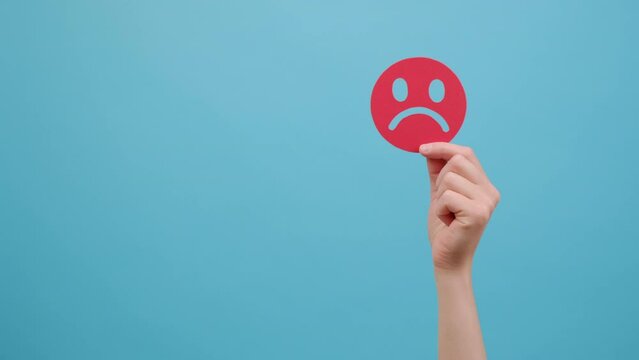 Close up of female hand holding red unsatisfied sad face emoticon, isolated on blue color background wall in studio with copy space for advertisement. Concept angry, stressed and negative emotion