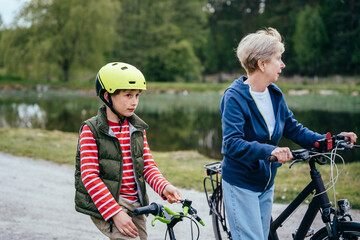 Naklejka premium Different generation family communication concept. Smiling senior woman with her grandson walking on a country lane with bicycles with lake on background.