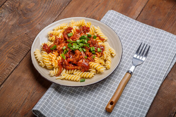 A plate with bolognese pasta on a wooden table next to a light napkin and a fork.