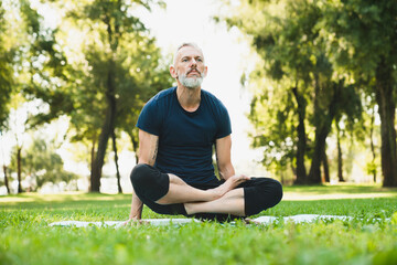 Caucasian mature male yogi sitting in lotus position on fitness mat on green grass in local park forest outdoors. Healthy lifestyle.