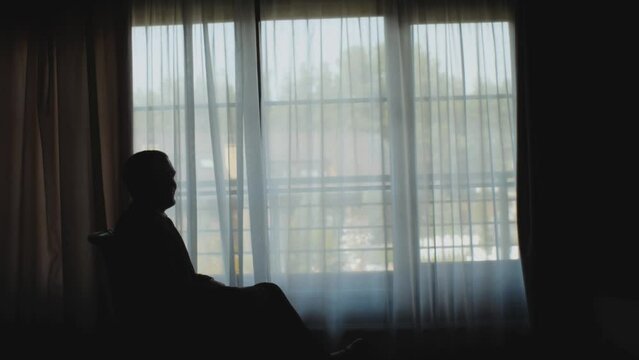 Silhouette of man walking from a dark room to the window. Guy in classic suit sit in chair near the window slow motion.