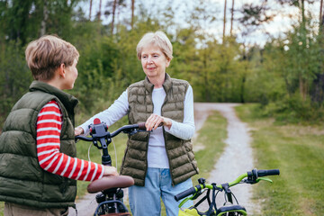 Young sportive slim short haired randmother teaches her grandson how to ride a bike at nature.