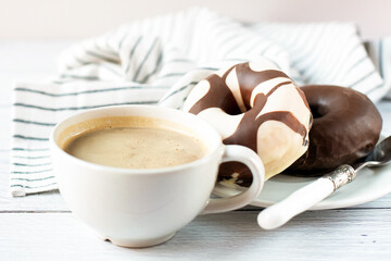 A cup of coffee, two chocolate donuts and a striped kitchen towel are on a light background. Theme is sweet delicious breakfast.