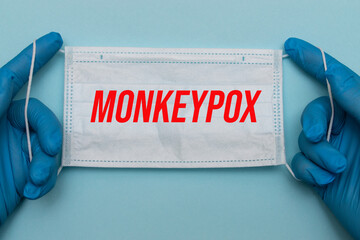 Hands in blue medical gloves hold a protective medical mask with the red inscription MONKEYPOX....