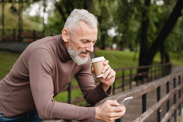 Handsome mature caucasian man using smartphone, calling his friends, colleagues, wife with good connection, texting online, surfing social media drinking coffee outdoors in park forest.