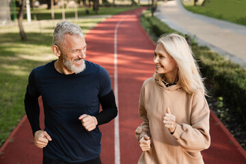 Senior mature couple running together in the park stadium looking at each other while jogging...