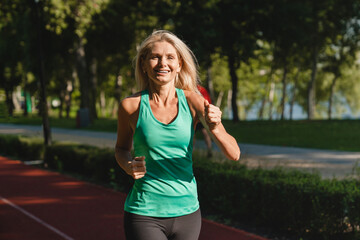 Happy mature caucasian woman running jogging in the morning, doing physical activities for slimming. Yoga workout outdoors in stadium.