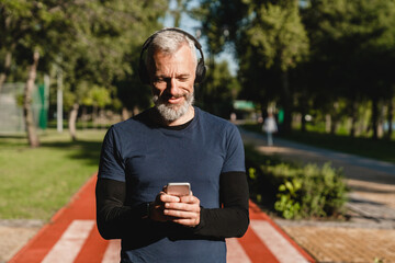 Naklejka na ściany i meble Closeup cropped portrait of a male caucasian mature athlete sportsman runner choosing sound track on smart phone listening to the music while jogging on stadium in the morning.