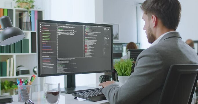 
Dolly out Over the Shoulder Shot of Young Businessman Sitting at Desk Using Desktop Computer Writing Program Code. Bright Office with coworkers. Caucasian Male Programmer or Software Engineer.