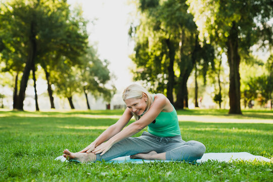 Caucasian mature pretty slim woman in sporty outfit stretching her leg on fitness mat during yoga class in park outdoors.