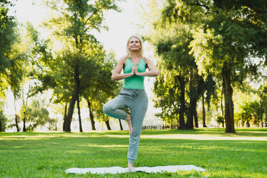 Balance. Caucasian mature woman standing on one foot meditating on fitness mat, feeling zen-like in public park. Healthy lifestyle