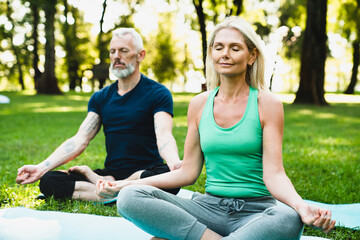 Zen-like lifestyle. Two mature people meditating in lotus position on fitness mats in public park together. Mature couple doing workout outdoors.
