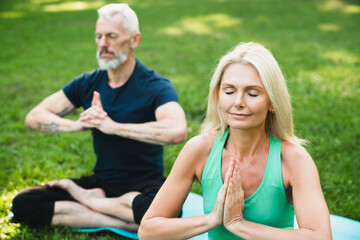 Mature couple relaxing meditating in lotus position together feeling zen-like on fitness mat in forest park. Active seniors, healthy lifestyle. Yoga class outdoors.