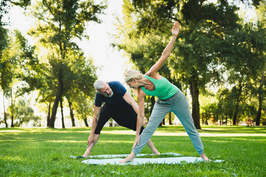 Caucasian fit healthy mature couple training together on fitness mat outdoors. Stretching exercises, yoga workout in public park with instructor coach.