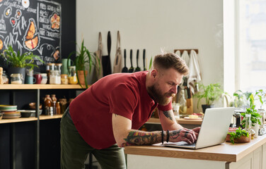 Young adult bearded Caucasian man with tattoos on arms surfing Internet on laptop in loft kitchen...