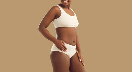 Woman is confident in her beauty and enjoys her size and type of figure standing on beige background. Cropped image of black woman in white cotton underwear who has no complexes about her appearance.