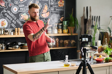 Stylish Caucasian food blogger making content for vlog dusting flour in air while preparing table...