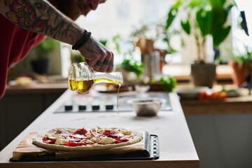 Young bearded man cooking pizza in kitchen at home adding some olive oil over it before putting it into oven