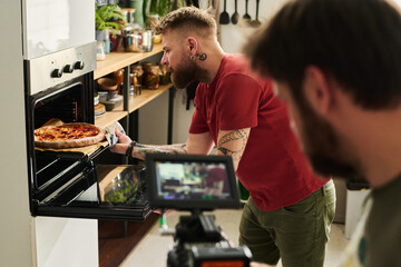 Unrecognizable cameraman filming modern bearded Caucasian man taking pizza out of hot oven