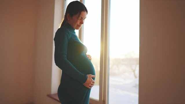 Woman Pregnant. Motherhood A Pregnancy Concept. 40 Year Old Pregnant Woman Stands In A Dress By The Window Holding Her Stomach Sunset Silhouette. Silhouette Of A Girl In By The Window