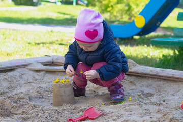 Baby girl caucasian plays with sand in the sandbox in spring or autumn, there is a sand castle