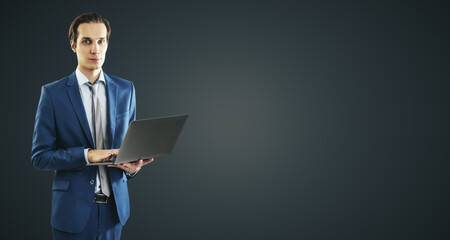 Young businessman in blue suit working with modern laptop, isolated on dark grey wall background, with blank place for you text or logo, mockup