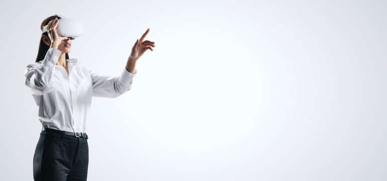 Young Businesswoman Working In Virtual Reality Environment In VR Glasses, Presses By Her Finger On Virtual Interface On Abstract White Backdrop. Innovate And Future Technology Concept, Mock Up