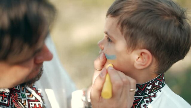 Dad draws yellow-blue flag of Ukraine on his son's cheek. Body art for support of Motherland. Family in vyshyvanka shirts. Patriotism, self-identity, love for native land