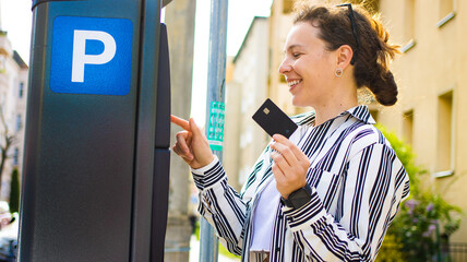 Happy elegant young woman pay for ticket in parking meter, holding credit card in hand,press button,standing near terminal on street outdoors.Using electronic payment,doing online banking transaction
