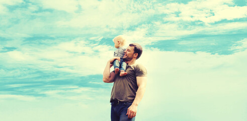 Happy strong father holding his son child on shoulder outdoors on blue sky background with clouds