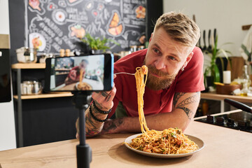 Food blogger eating homemade pasta while filming recipe and mukbang with smartphone on tripod in...