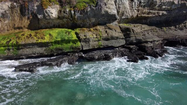 aerial footage of the vast blue ocean water with waves crashing into the rocks coved with lush green algae with a gorgeous clear blue sky at West Street Beach in Laguna Beach California USA