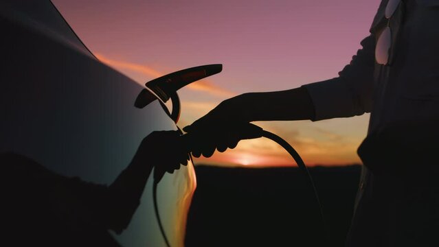 Scenic pink purple sunset with golden clouds on background of zero emission modern electric car. Woman silhouette is charging eco friendly. Slow motion RED camera shot futuristic innovative technology