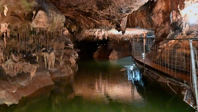 Water in cave with lights reflecting off formations of limestone and calcite on walls, with elevated pathway for people to walk with guide on tours.